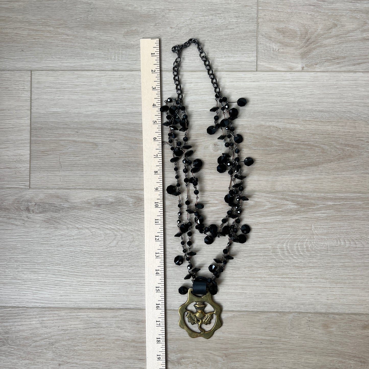 Black beaded necklace with a brass medallion on a wooden surface next to a ruler for scale.