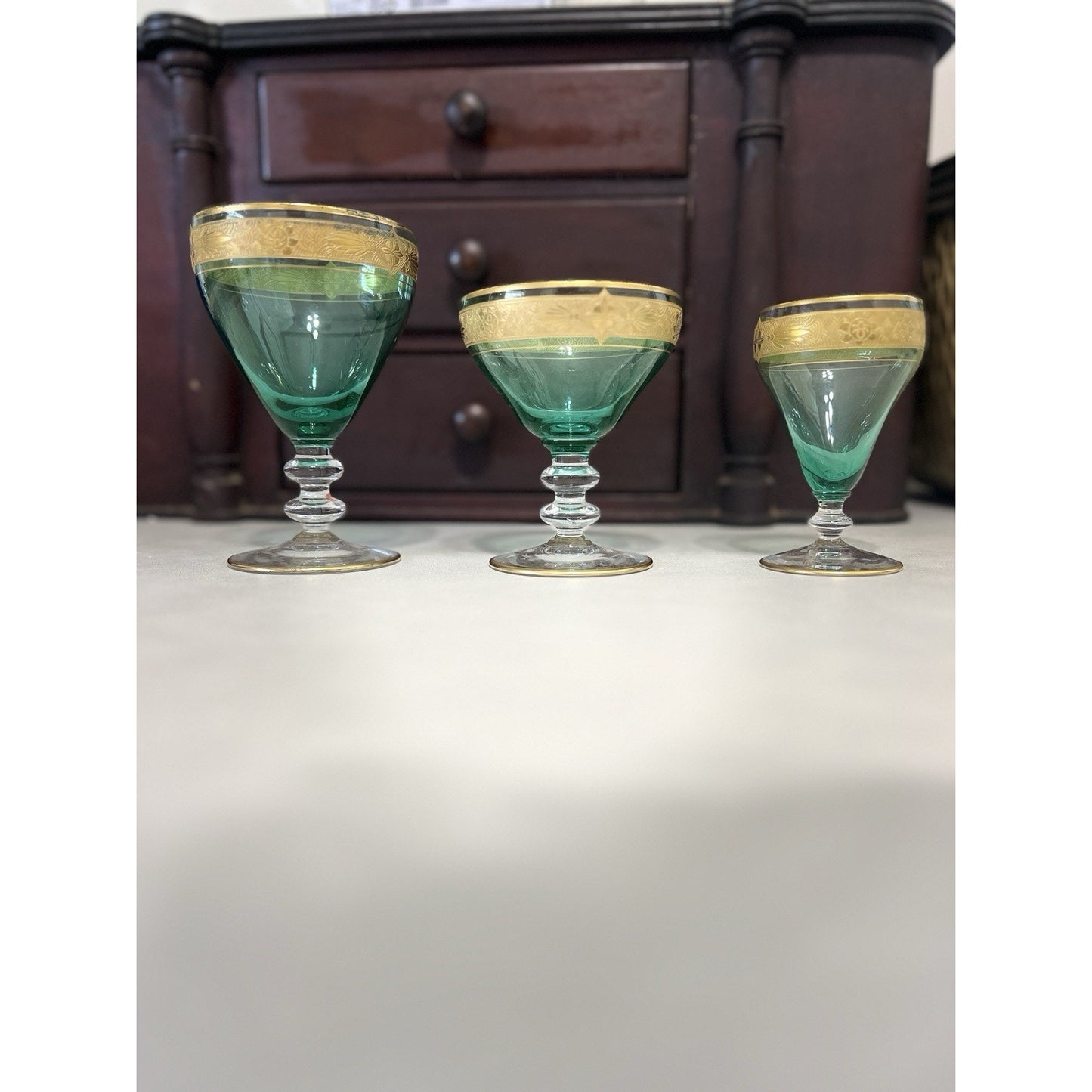 Three vintage green glass coupe glasses with gold rims lined up on a white surface in front of a dark wooden chest of drawers.