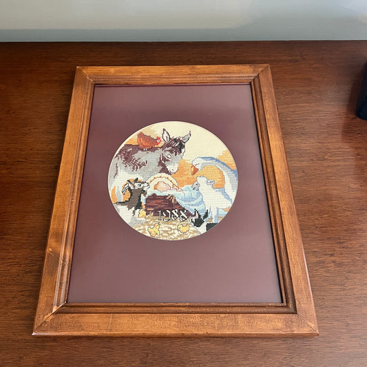A framed circular needlepoint canvas featuring a rustic scene with a donkey, sheep, and a basket, mounted on a dark brown mat board inside a medium brown wooden frame, resting on a wooden table.