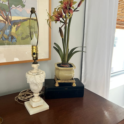 Decorative lamp and potted plant on a wooden surface with a painting in the background.