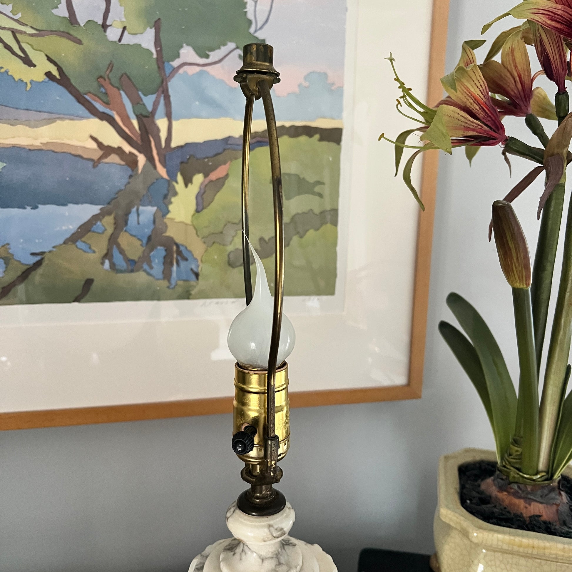 Decorative lamp with a white base and brass fitting, next to a potted plant and framed artwork.