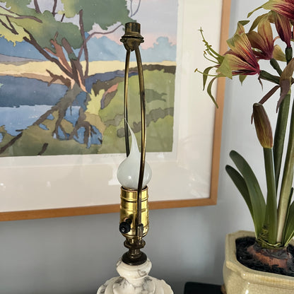 Decorative lamp with a white base and brass fitting, next to a potted plant and framed artwork.