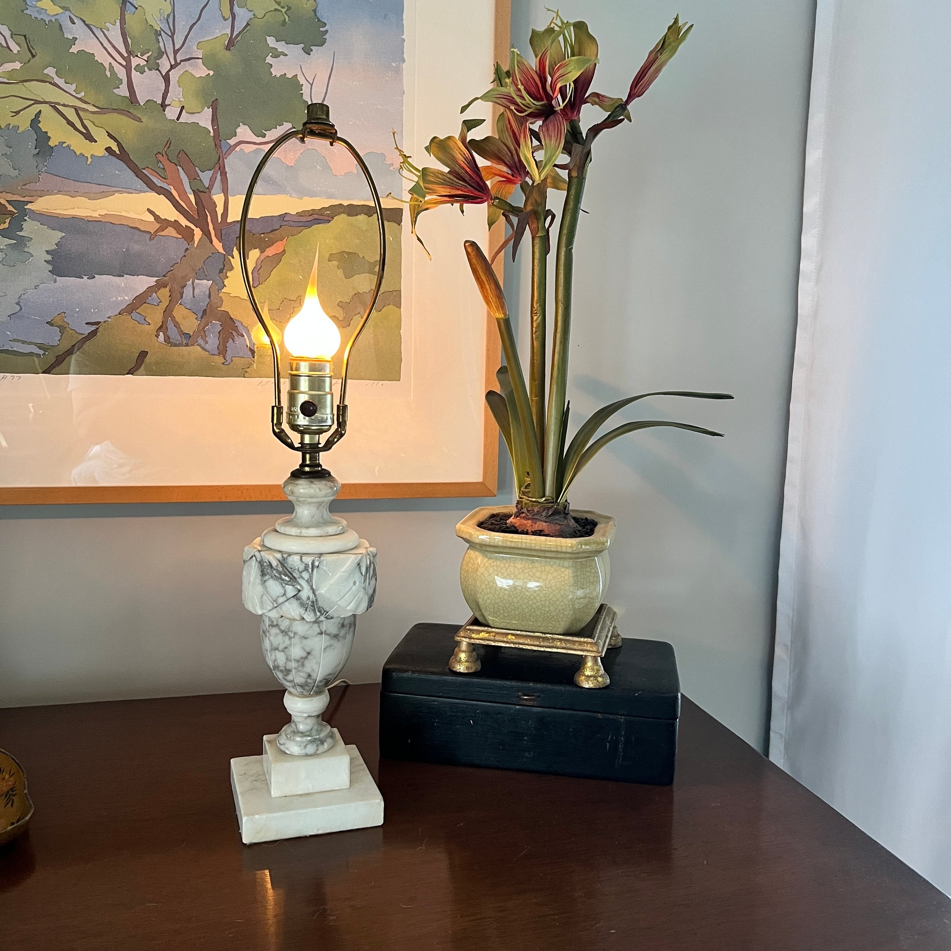 Marble lamp with a light bulb and a potted plant on a wooden surface, with a painting in the background.