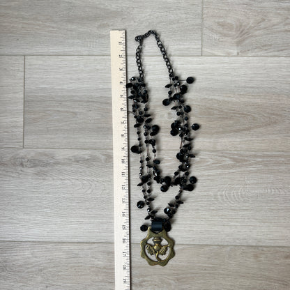 Black beaded necklace with a brass medallion on a wooden surface next to a ruler for scale.