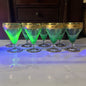 A set of six clear glass coupe-style cocktail glasses with gold bands around the rims, green from uv light arranged on a white marble surface with blue ambient lighting.
