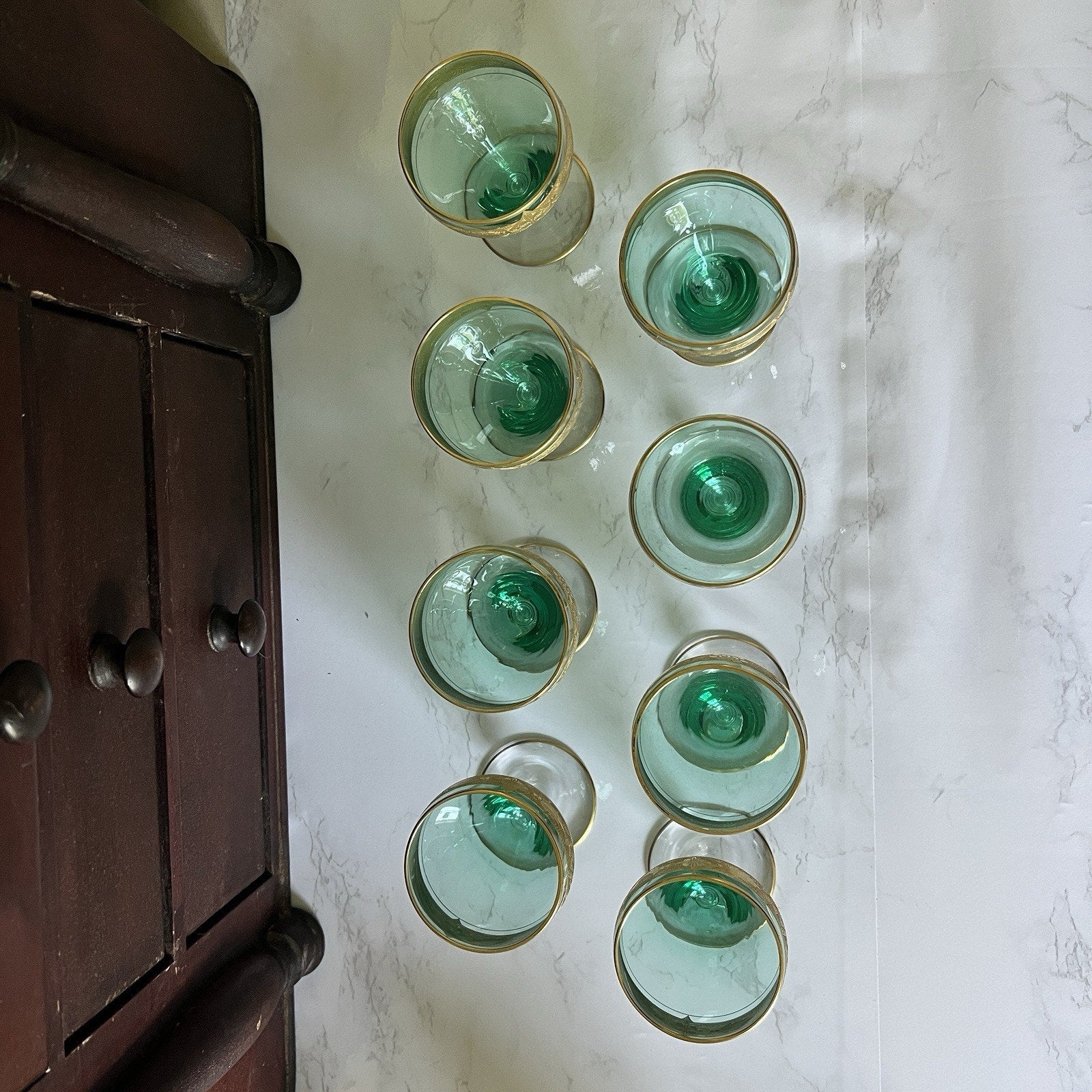Eight vintage green-tinted glass tumblers with gold rims arranged on a white marble surface next to a dark brown wooden cabinet.