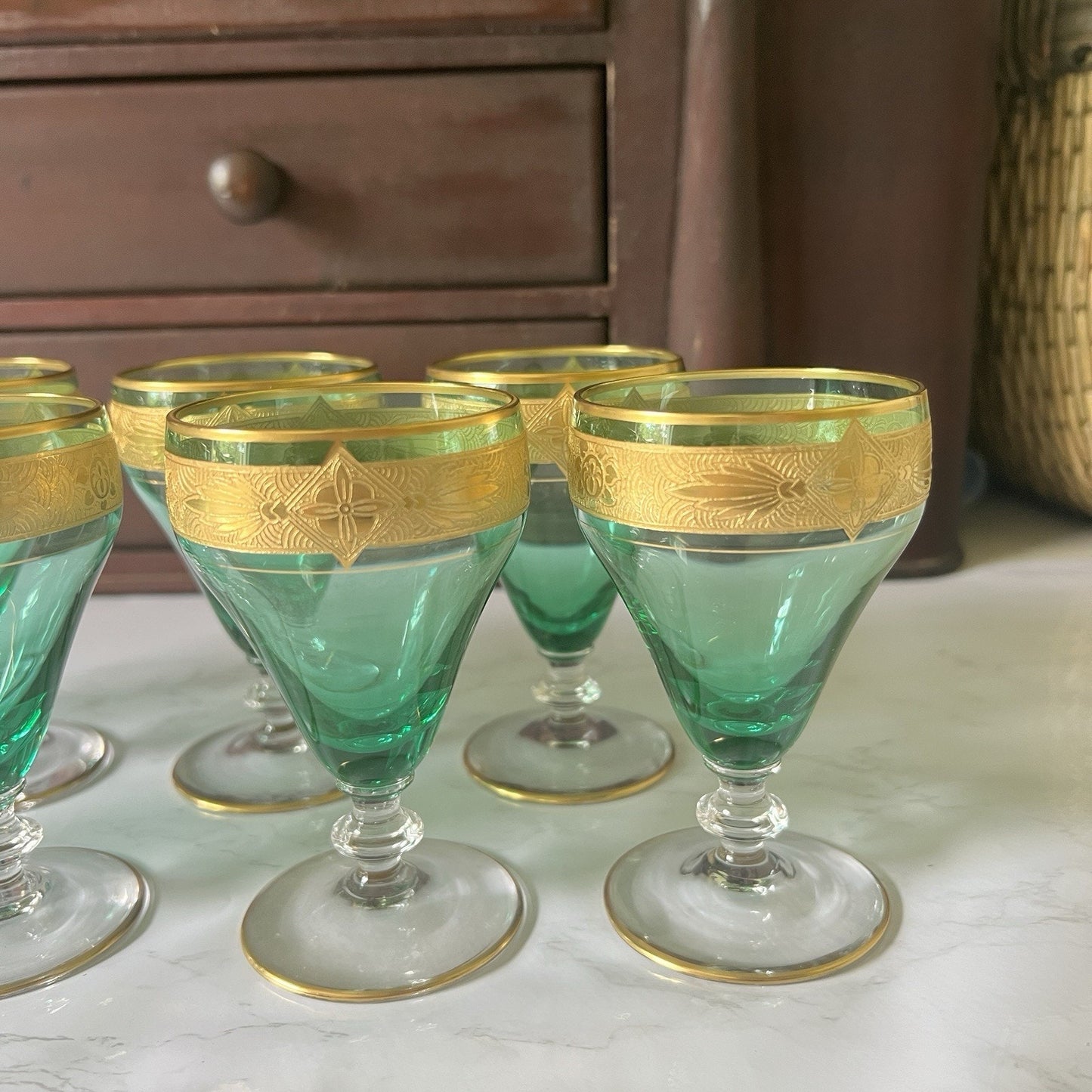 A set of vintage green glass goblets with ornate gold bands near the rim, arranged on a light marble surface with a dark wooden cabinet in the background.