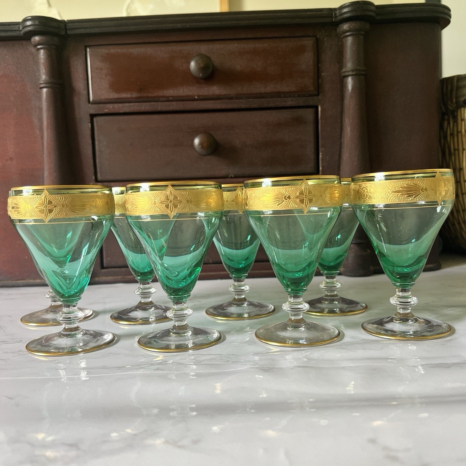 Six green crystal glasses with gold-trimmed rims arranged on a white marble surface in front of a dark wooden cabinet.