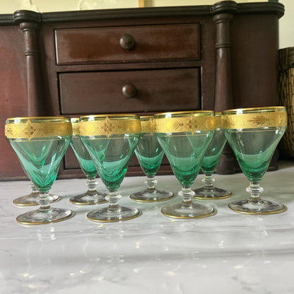 Six green crystal glasses with gold-trimmed rims arranged on a white marble surface in front of a dark wooden cabinet.