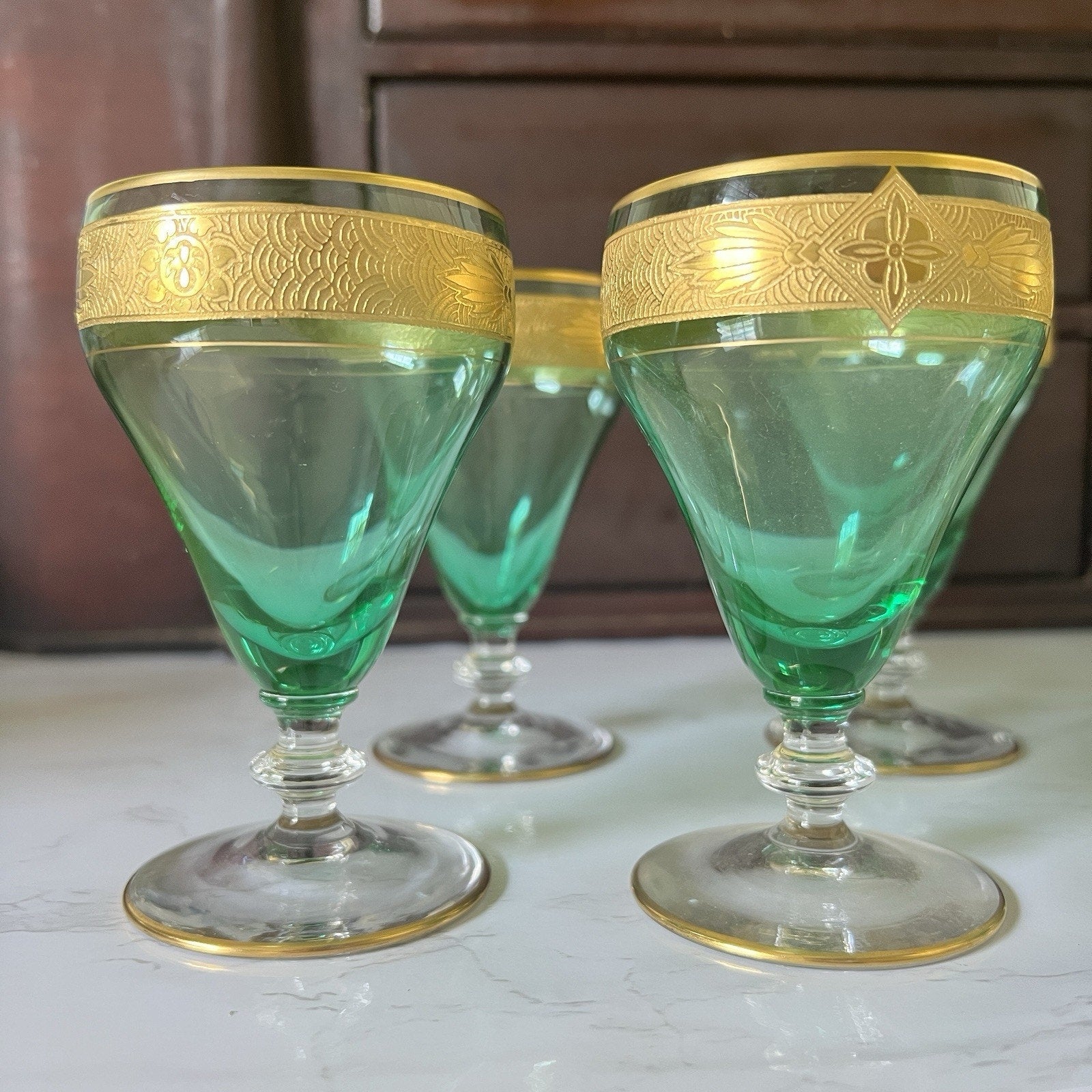 Three vintage green glass goblets with gold-trimmed bands and gold-rimmed edges, arranged on a light marble surface with a dark wooden cabinet in the background.