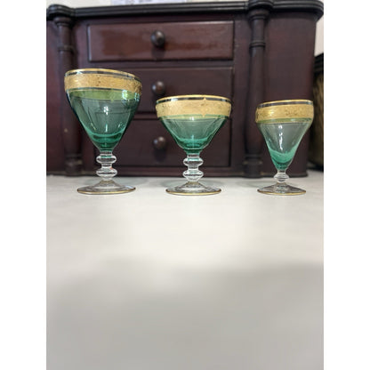 Three vintage green glass coupe glasses with gold rims lined up on a white surface in front of a dark wooden chest of drawers.