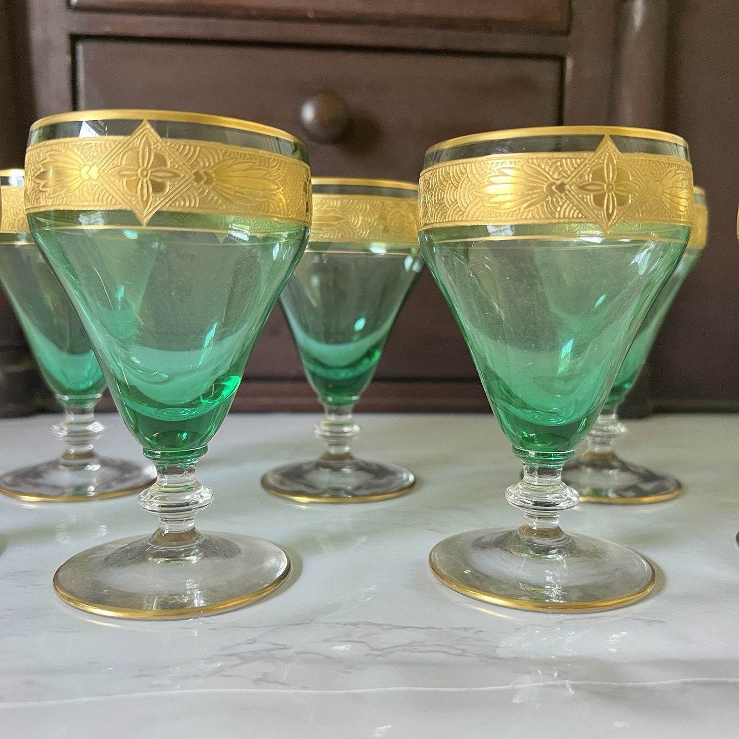 A set of green glass goblets with ornate gold bands around the rims, displayed on a light marble surface with a dark wooden cabinet in the background.