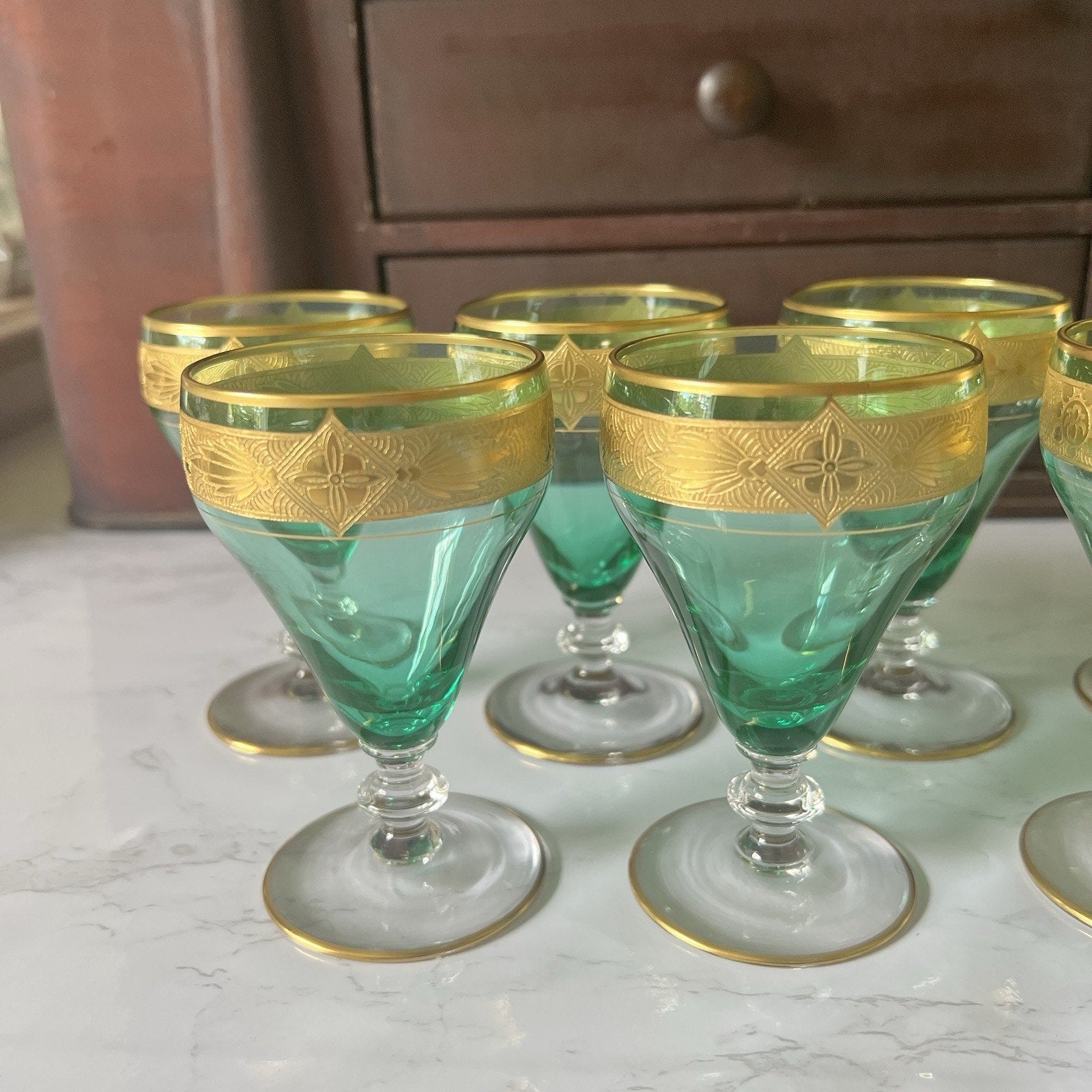 A set of vintage green glass coupe glasses with gold trim, displayed on a white marble surface with a wooden cabinet in the background.