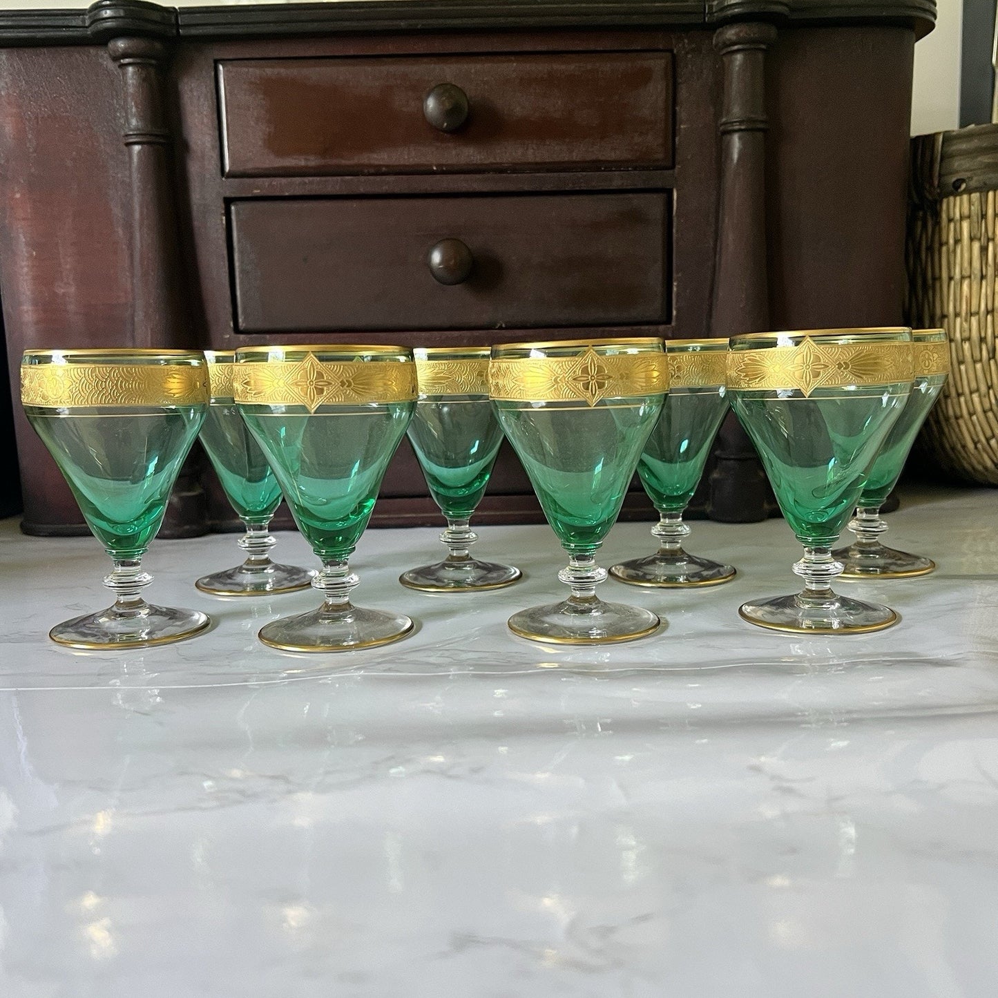 A set of eight green-tinted glass cordial glassed with gold-trimmed rims, arranged on a white marble surface in front of a dark wooden chest of drawers.