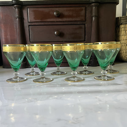 A set of eight green-tinted glass cordial glassed with gold-trimmed rims, arranged on a white marble surface in front of a dark wooden chest of drawers.