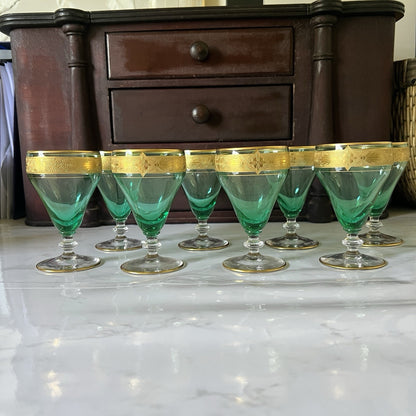 A set of eight green-tinted glass cordial glasses with gold bands, arranged on a white marble countertop in front of a dark wooden chest of drawers.