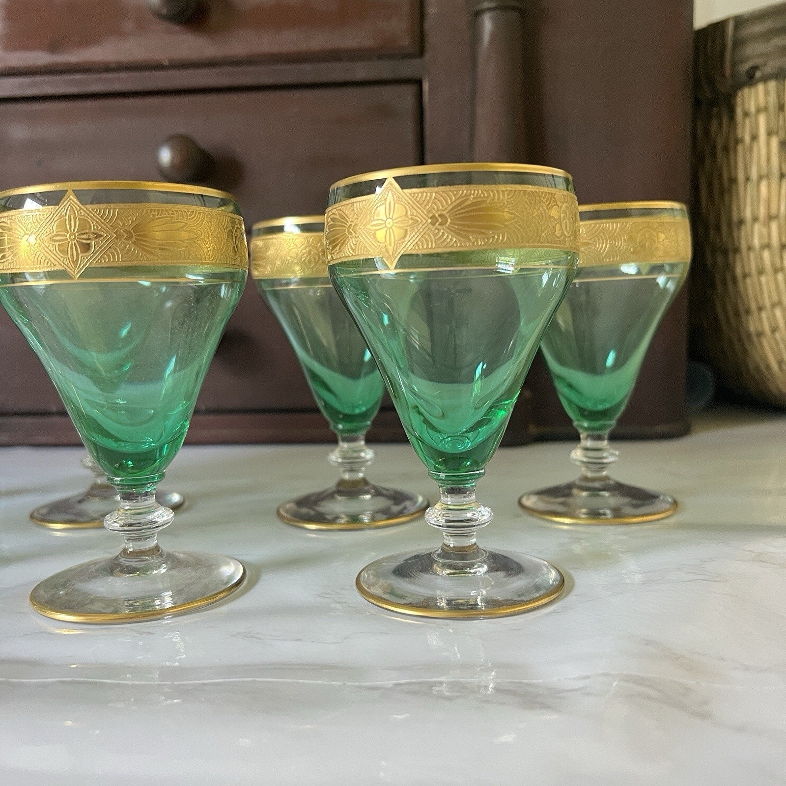 Five green glass coupe glasses with gold etched borders, arranged on a white marble surface with a dark wooden cabinet in the background.