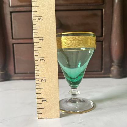 A small green-tinted glass with a gold patterned rim and gold base trim, shown next to a wooden ruler for scale on a light surface with a dark wooden cabinet in the background.