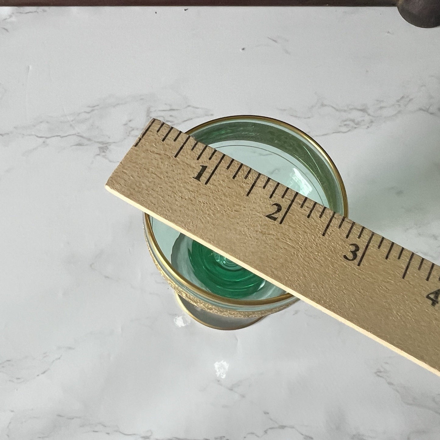 A  green glass with a  gold-toned rim, shown on a white marble surface with a wooden ruler placed diagonally across it for scale.
