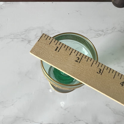 A  green glass with a  gold-toned rim, shown on a white marble surface with a wooden ruler placed diagonally across it for scale.