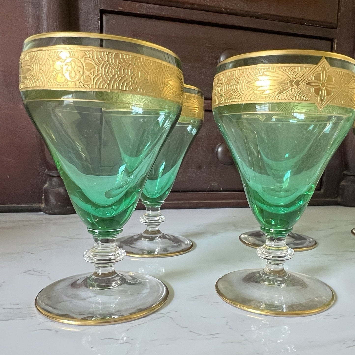Four green glass cordial glasses with ornate gold bands around the rims, displayed on a light marble surface with a dark wooden cabinet in the background.