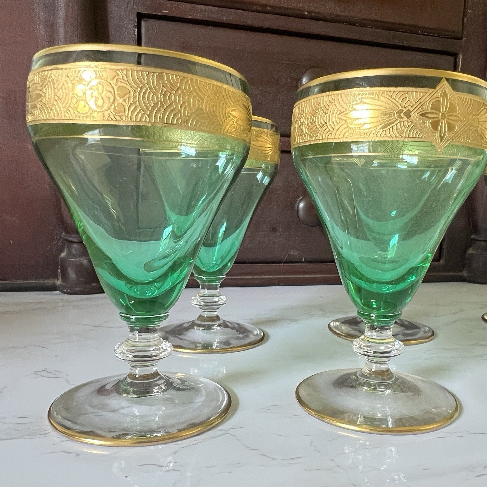 Four green glass cordial glasses with ornate gold bands around the rims, displayed on a light marble surface with a dark wooden cabinet in the background.