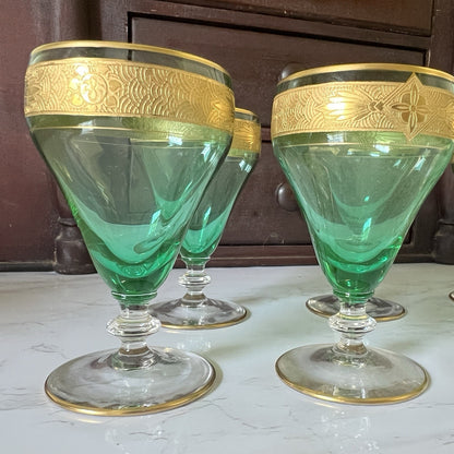 Four green glass cordial glasses with ornate gold bands around the rims, displayed on a light marble surface with a dark wooden cabinet in the background.