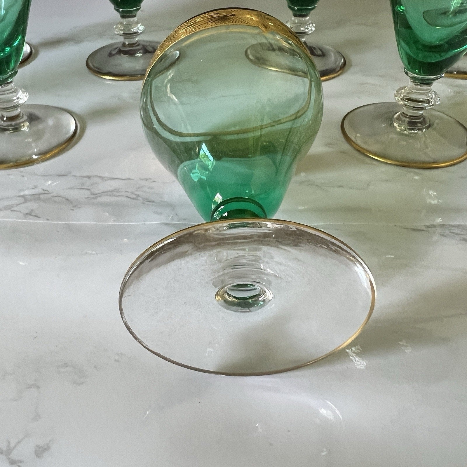 A green glass goblet with a gold rim, shown lying on its side on a white marble surface, with other matching green and gold glasses partially visible in the background.