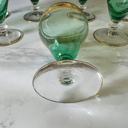 A green glass goblet with a gold rim, shown lying on its side on a white marble surface, with other matching green and gold glasses partially visible in the background.