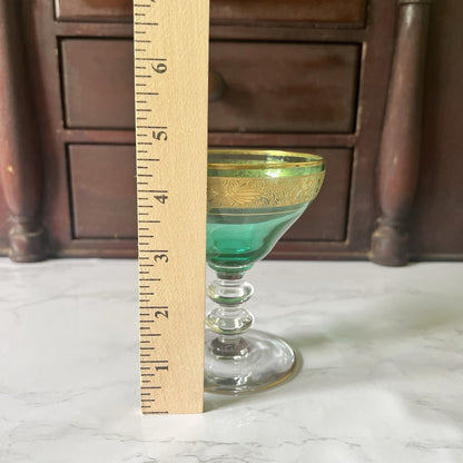 A small green glass coupe-style cup with a gold rim stands on a white marble surface, next to a wooden ruler showing measurements, with a dark wooden cabinet in the background.