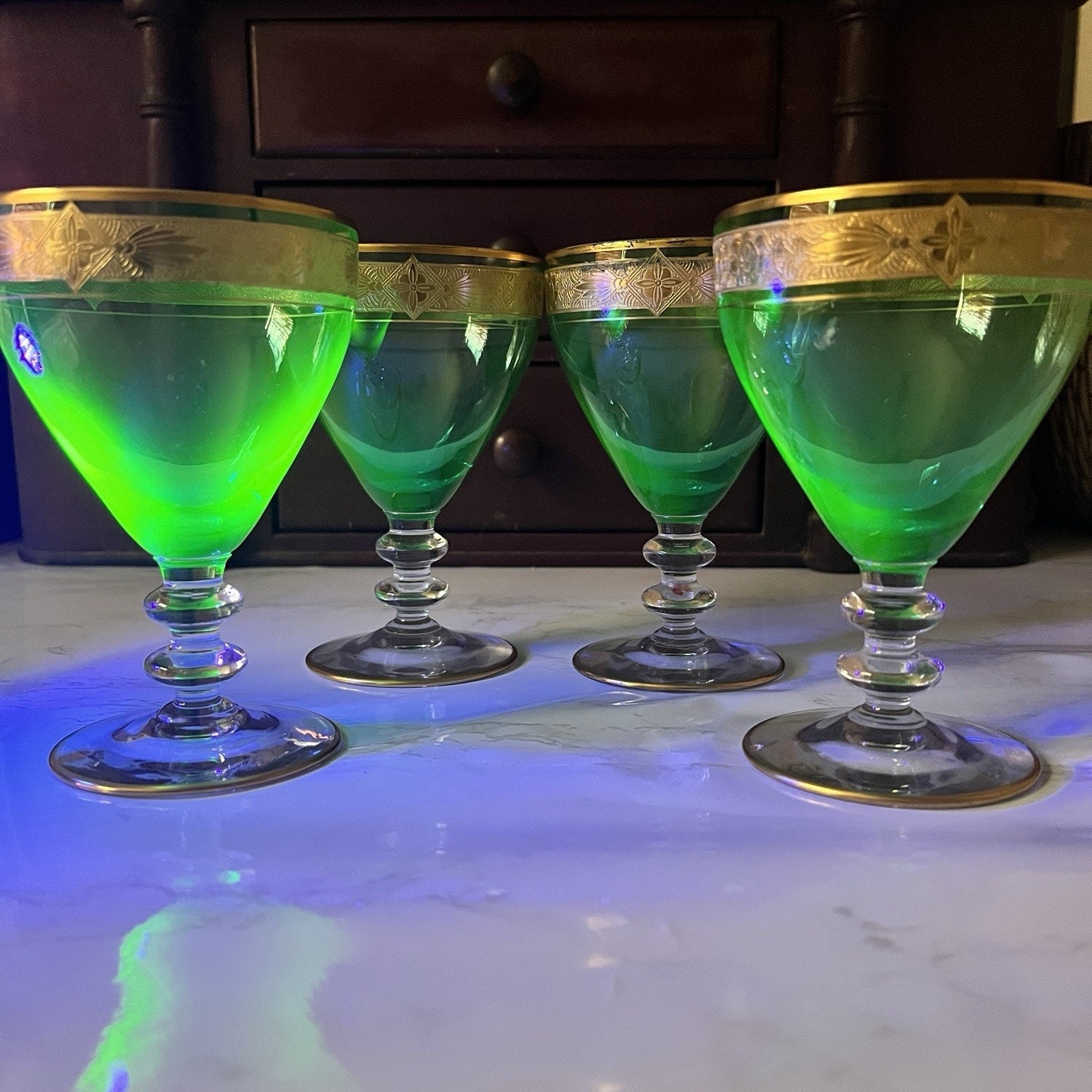 Four green cocktail glasses with gold-rimmed edges arranged on a white marble surface with blue and green reflections, set against a dark wooden cabinet background.