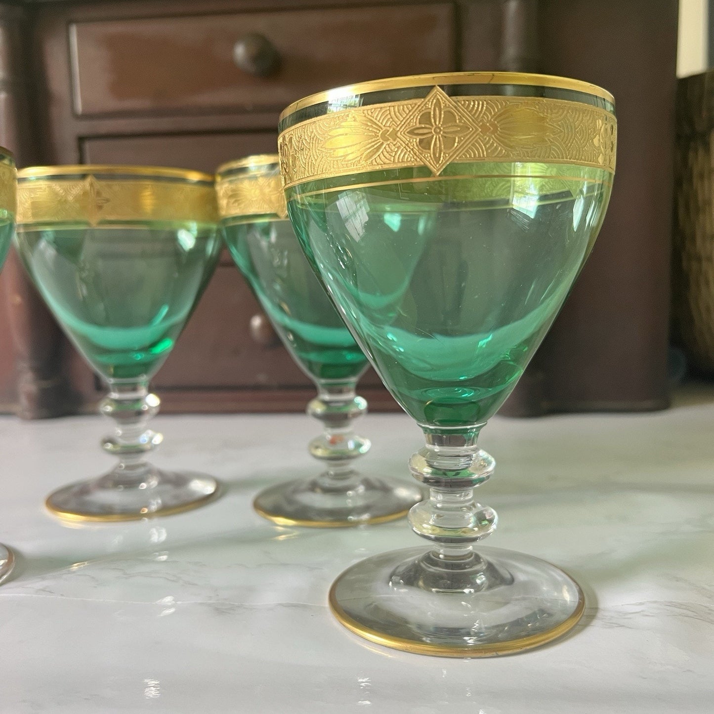 A set of green glass goblets with ornate gold bands around the rims, displayed on a light marble surface with a dark wooden cabinet in the background.