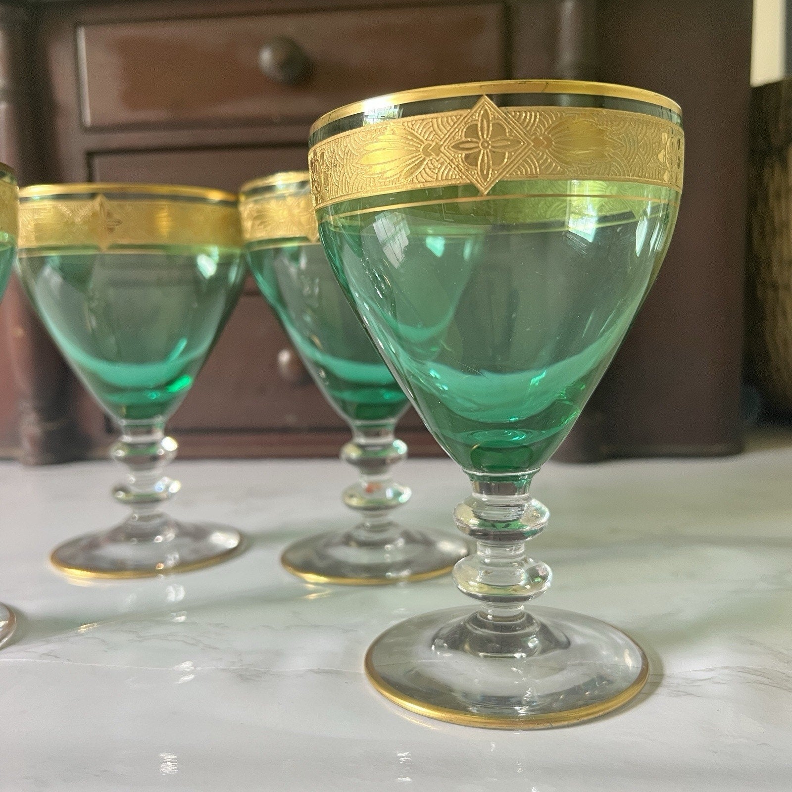 A set of green glass goblets with ornate gold bands around the rims, displayed on a light marble surface with a dark wooden cabinet in the background.