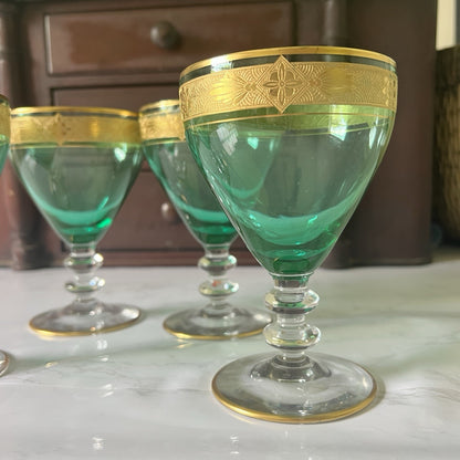 A set of green glass goblets with ornate gold bands around the rims, displayed on a light marble surface with a dark wooden cabinet in the background.