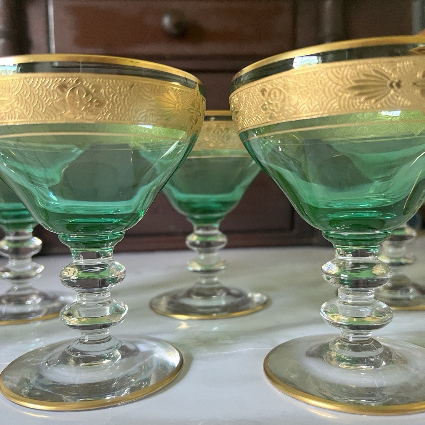 A set of green glass coupe-style goblets with ornate gold rims, displayed on a light surface with a dark wooden cabinet in the background.