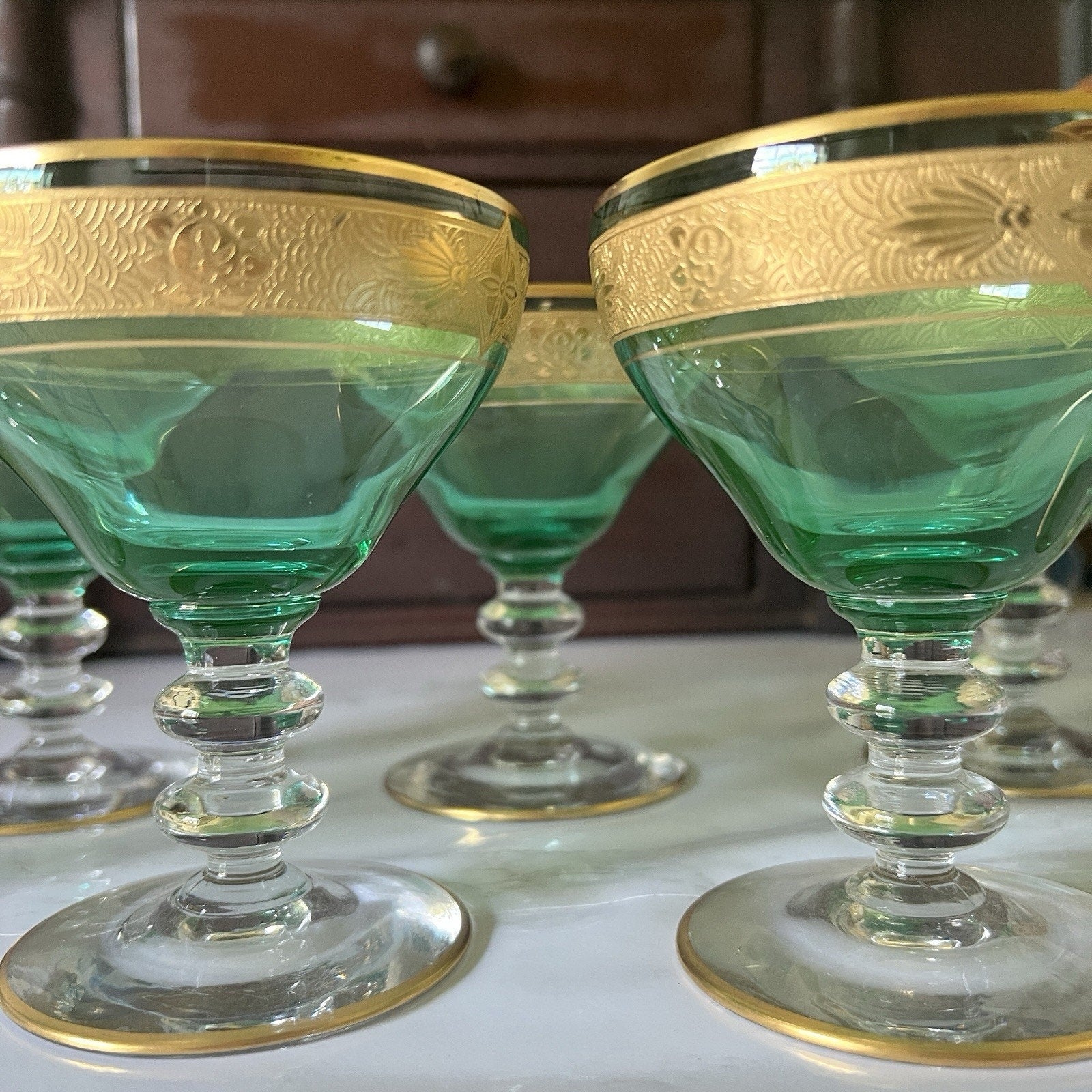 A set of green glass coupe-style goblets with ornate gold rims, displayed on a light surface with a dark wooden cabinet in the background.