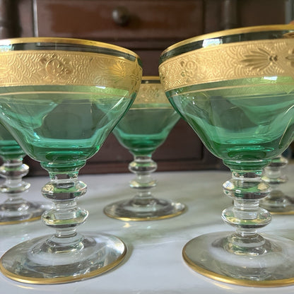 A set of green glass coupe-style goblets with ornate gold rims, displayed on a light surface with a dark wooden cabinet in the background.