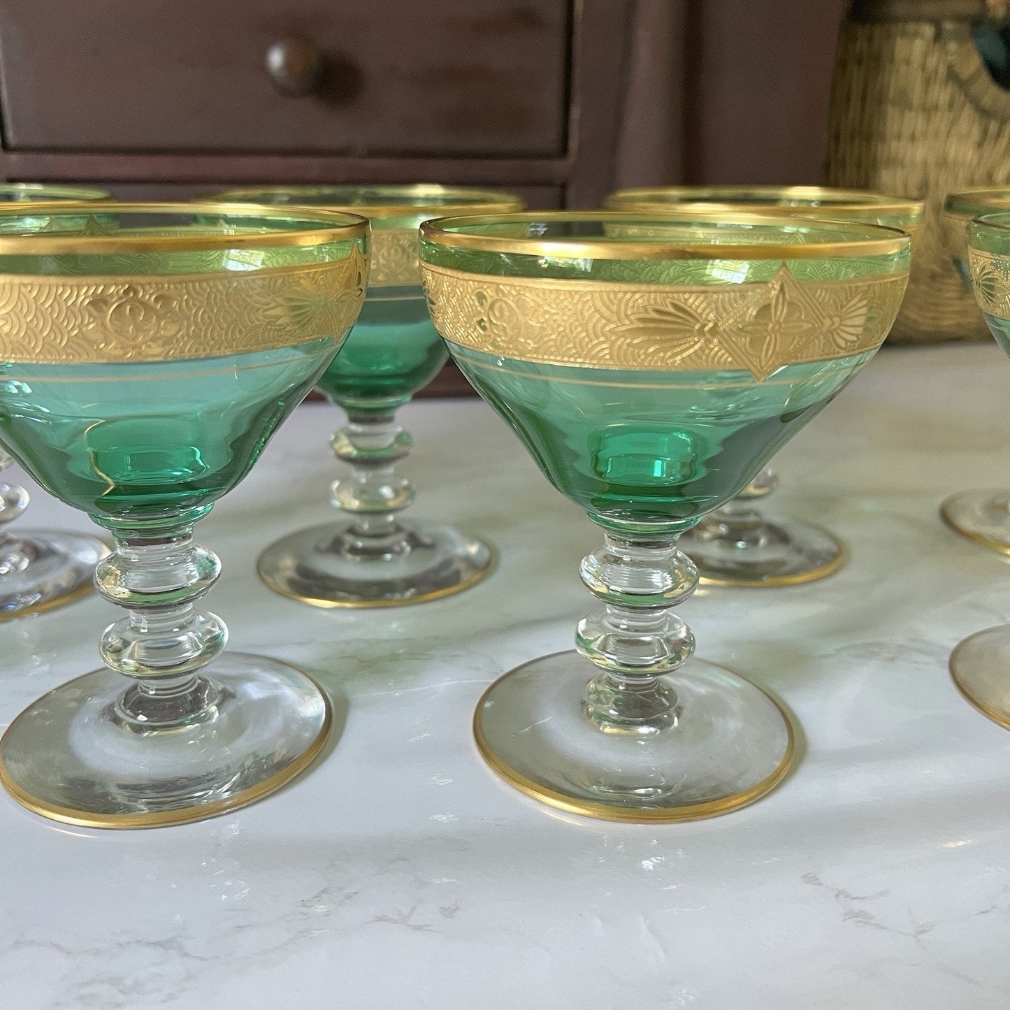 A set of green and gold coupe glasses arranged on a white marble surface with a dark wooden cabinet in the background.