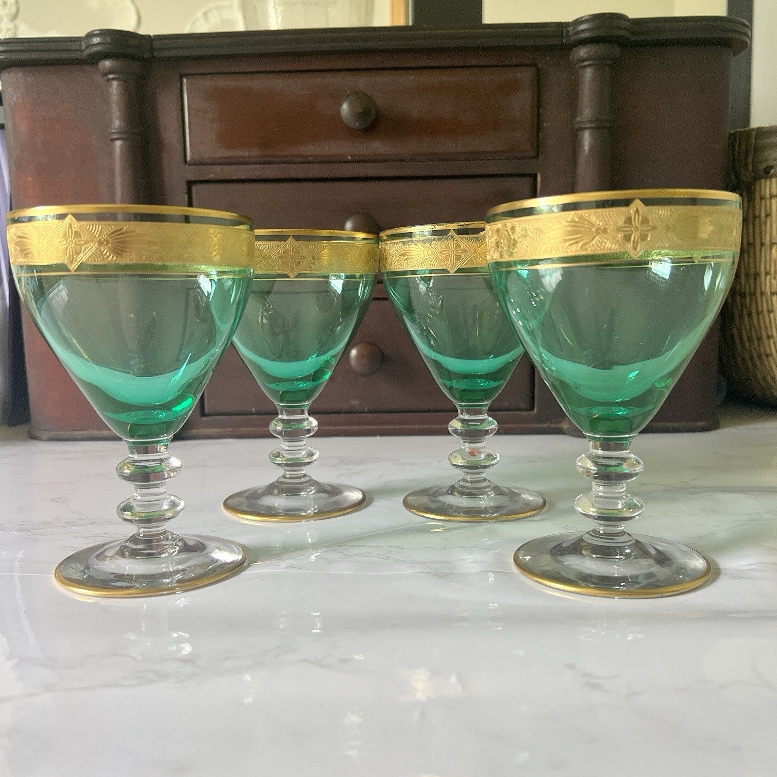 Four green-tinted glass coupe cups with gold decorative bands, arranged on a white marble surface with a dark wooden cabinet in the background.