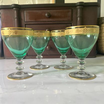 Four green-tinted glass coupe cups with gold decorative bands, arranged on a white marble surface with a dark wooden cabinet in the background.