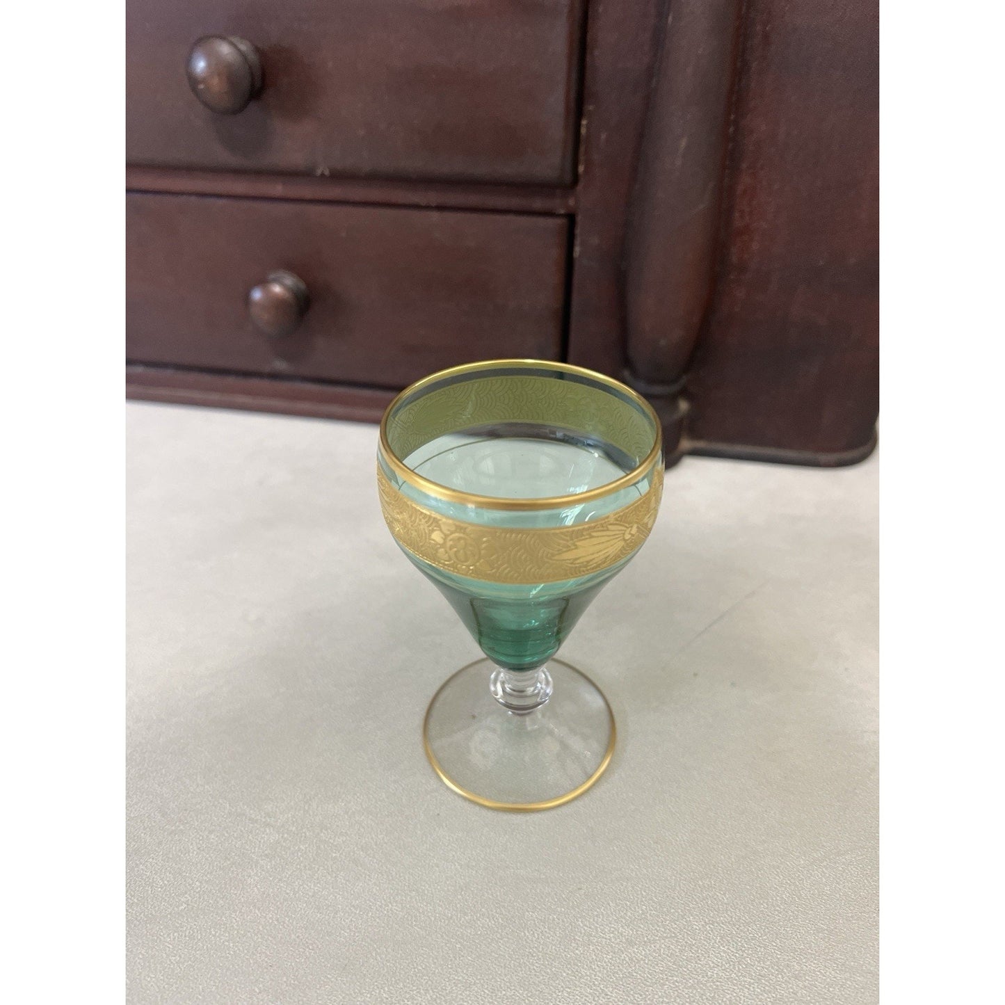 A vintage green glass goblet with a textured gold band around the upper half and a thin gold rim, set on a light gray floor in front of a dark wooden dresser.