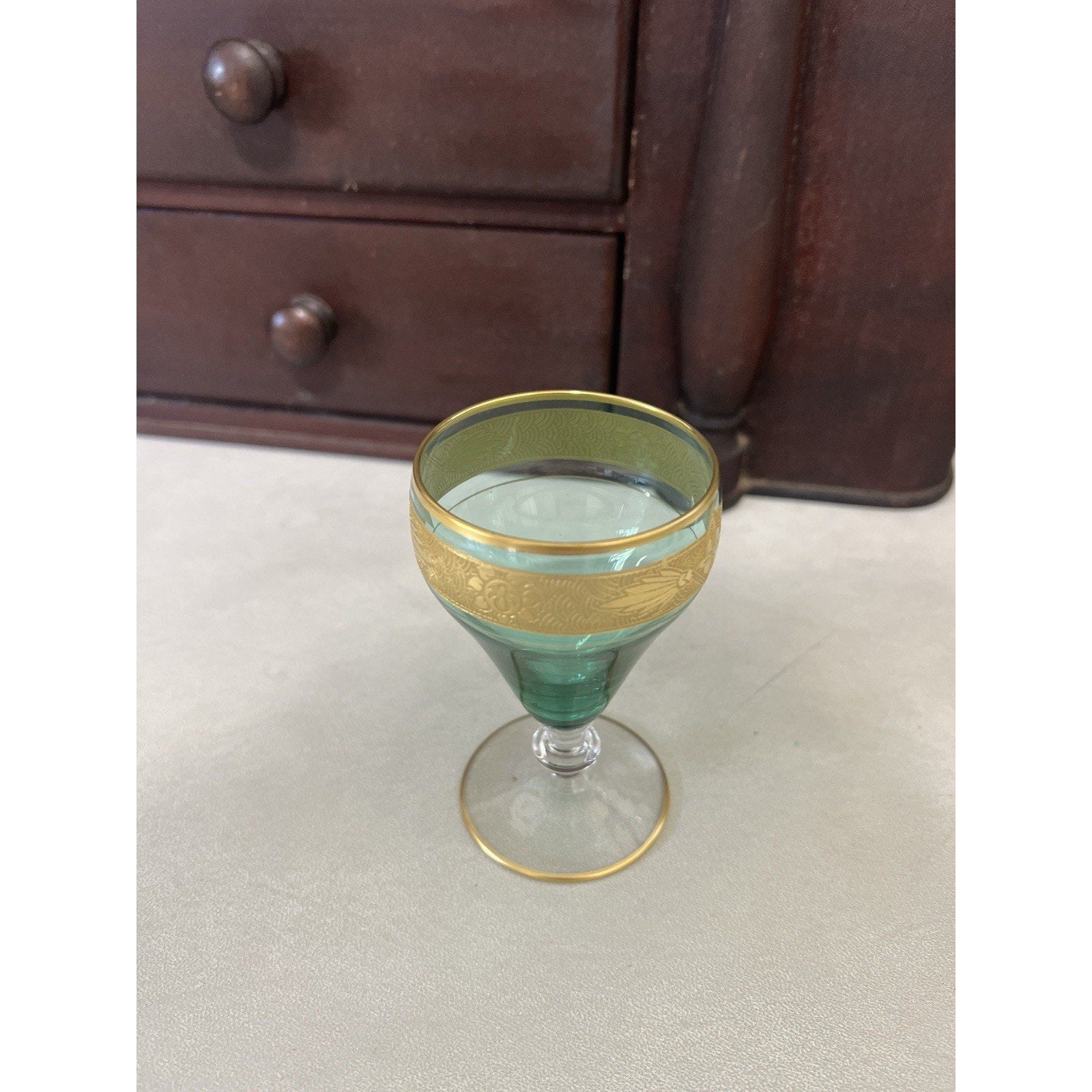A vintage green glass goblet with a textured gold band around the upper half and a thin gold rim, set on a light gray floor in front of a dark wooden dresser.