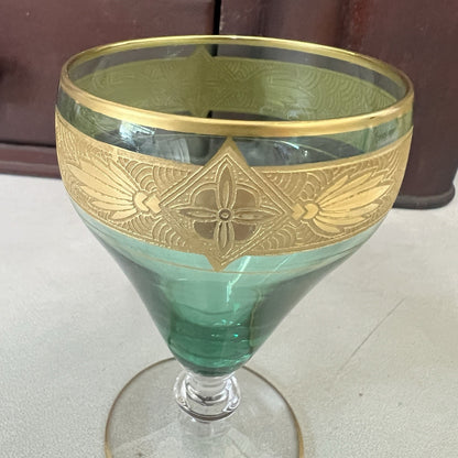 A green glass goblet with an ornate gold band featuring a central floral motif and leaf details, set against a neutral indoor background.