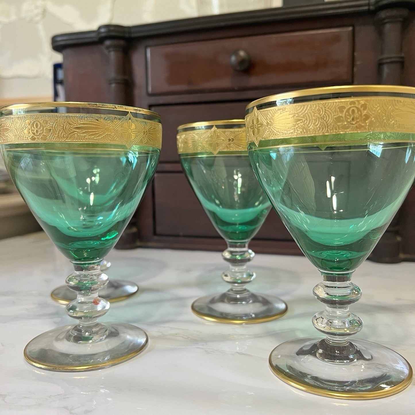 Three vintage green glass goblets with gold trim and ornate detailing on the rims, arranged on a light marble surface with a dark wooden cabinet in the background.