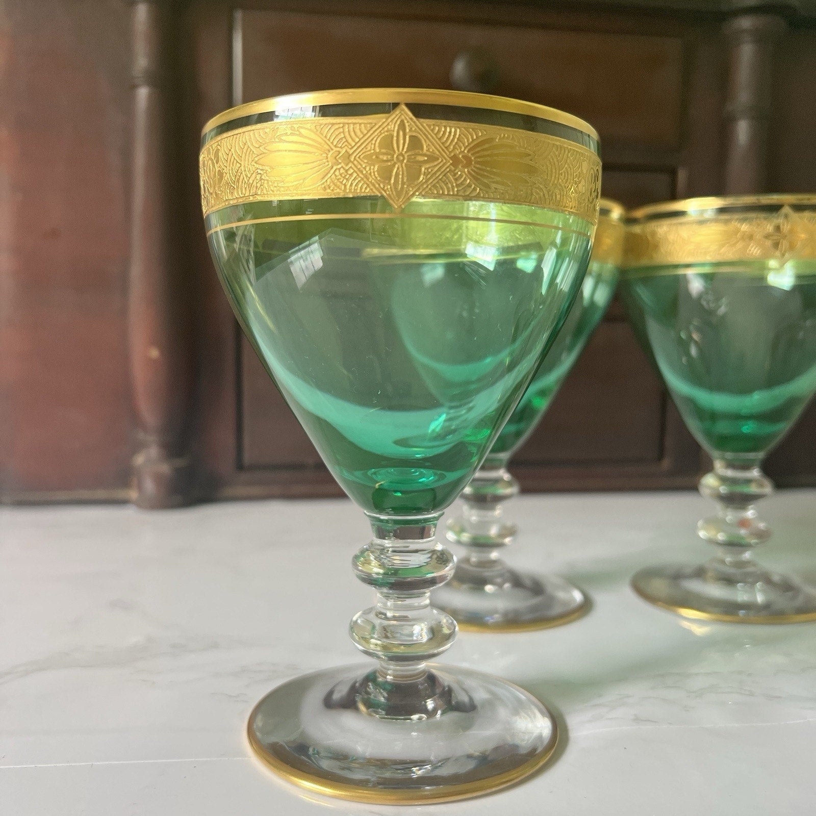 Three green glass goblets with ornate gold bands near the rim, displayed on a light surface with a dark wooden cabinet in the background.