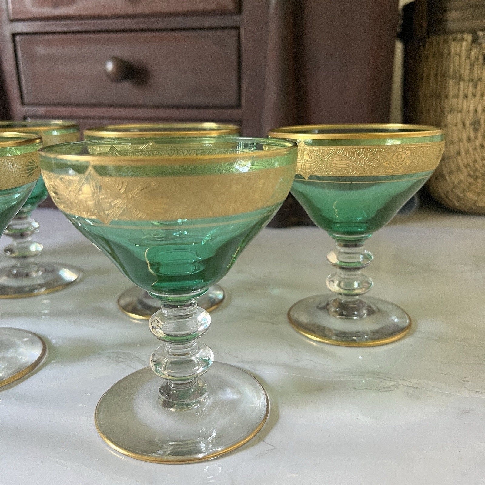 A set of green and gold coupe glasses with etched floral detailing, displayed on a light marble surface with a dark wooden cabinet in the background.