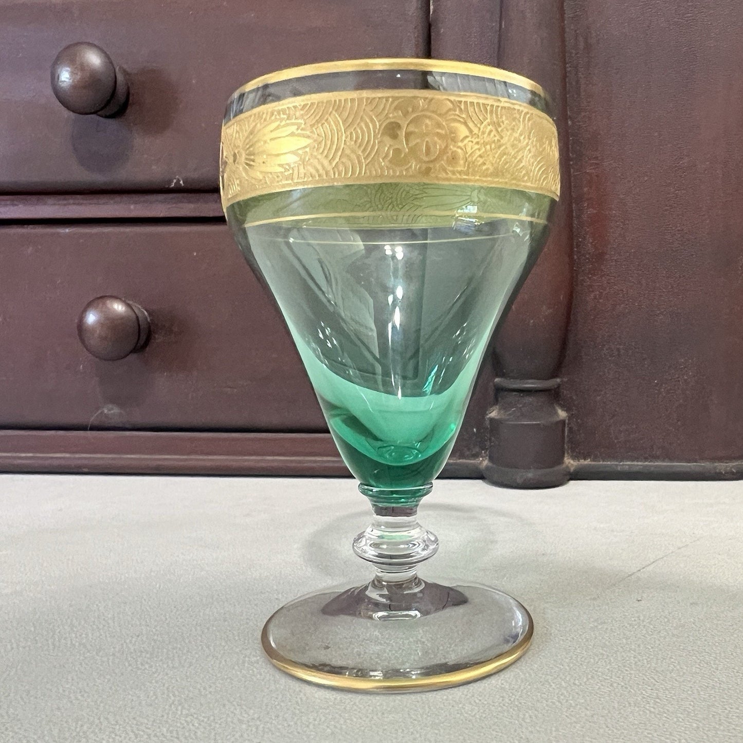 A vintage green glass goblet with a gold-colored decorative band around the rim, set against a dark wooden cabinet background.