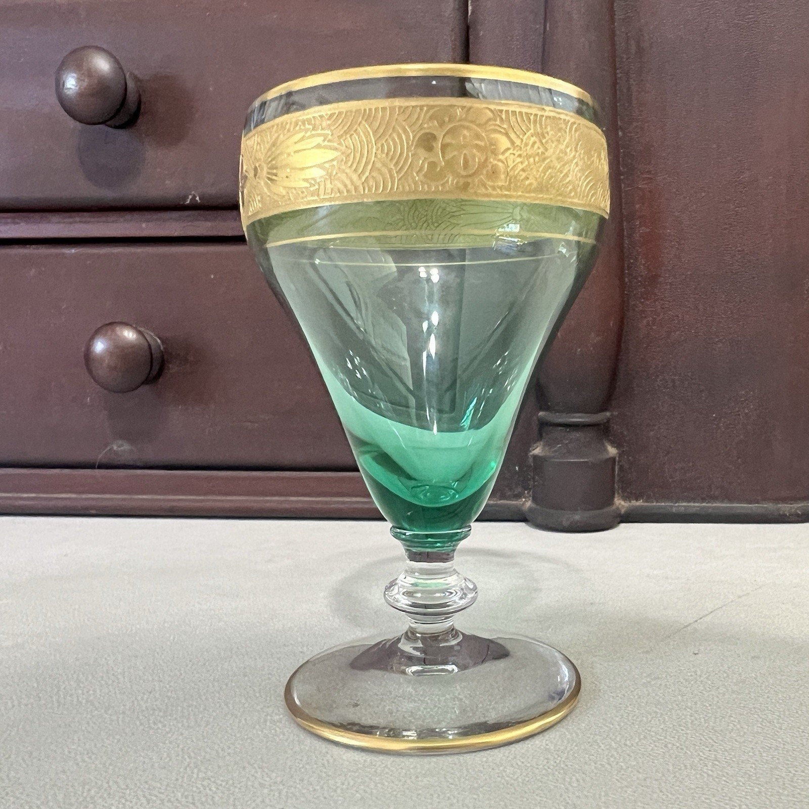 A vintage green glass goblet with a gold-colored decorative band around the rim, set against a dark wooden cabinet background.