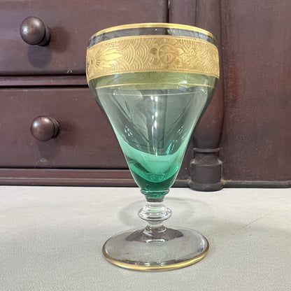 A vintage green glass goblet with a gold-colored decorative band around the rim, set against a dark wooden cabinet background.