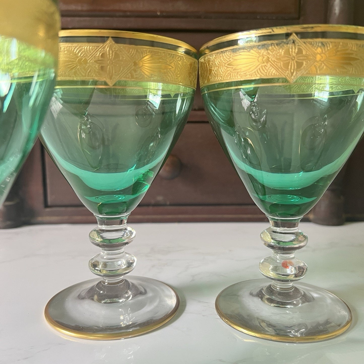 Two green glass goblets with gold trim and etched detailing on the rims, displayed on a white surface with a dark wooden background.